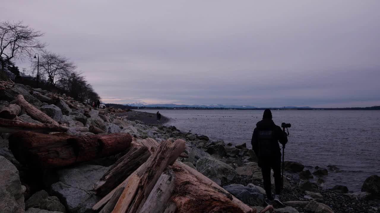 Un uomo cammina con treppiede e macchina fotografica su una spiaggia rocciosa e sabbiosa coperta da legni e massi, con persone che camminano in lontananza, nuvole, alberi e un parco di roccia bianca