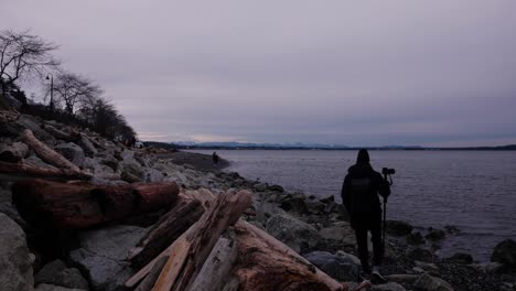 man walking with tripod and camera over sandy rock beach covered in drift wood and boulders with people walking in the distance clouds trees white rock park