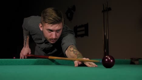 a brutal man with tattoos on his hands is playing russian billiards in a bar.