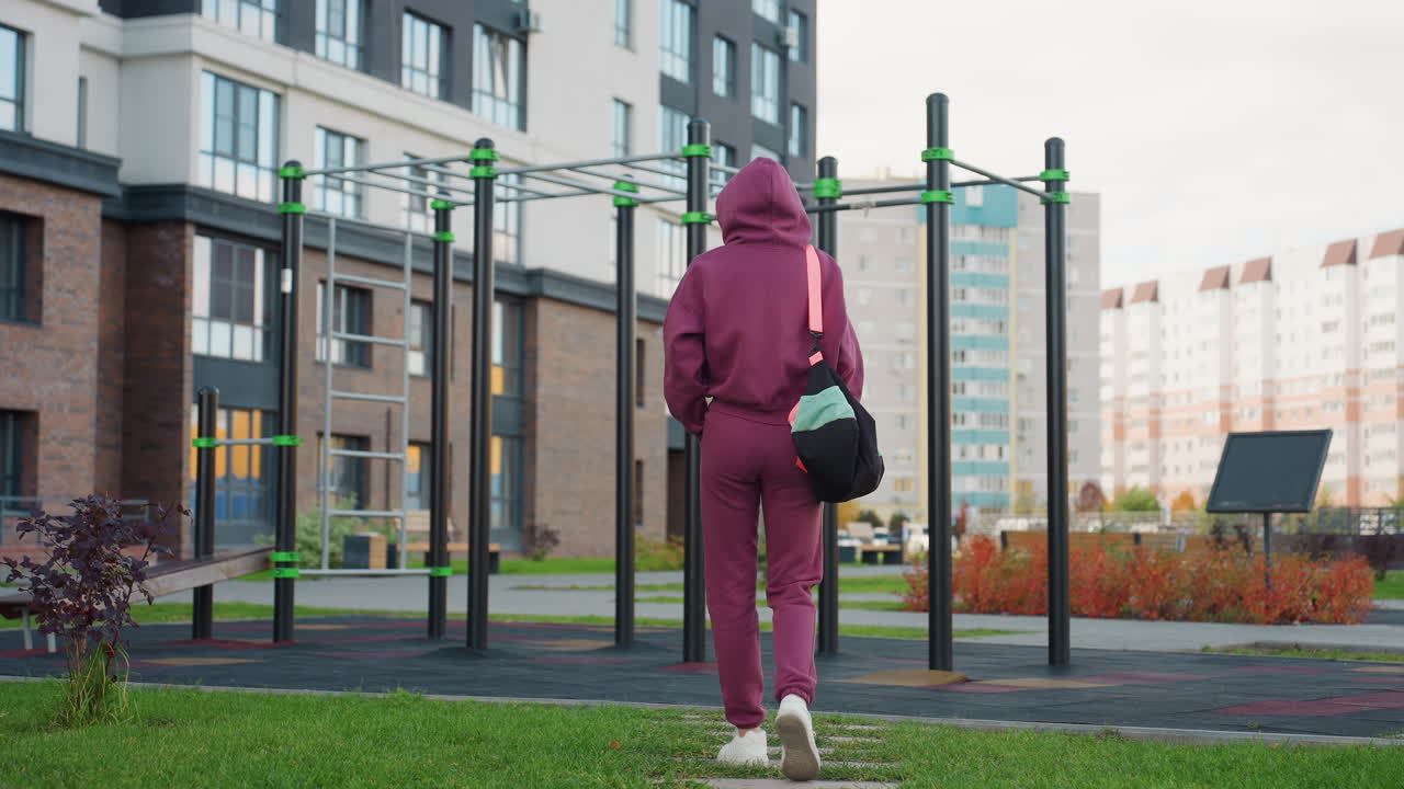 Workout enthusiast wearing hoodie walks with sport bag slung over shoulder, hands in pockets, approaching exercise bars in urban outdoor gym area framed by apartments, sunlight on confident form