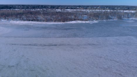 Pan-down-drone-shot-of-a-frozen-river-and-frozen-flooded-wood-while-the-drone-rises-in-the-air-getting-a-wider-view-of-the-river