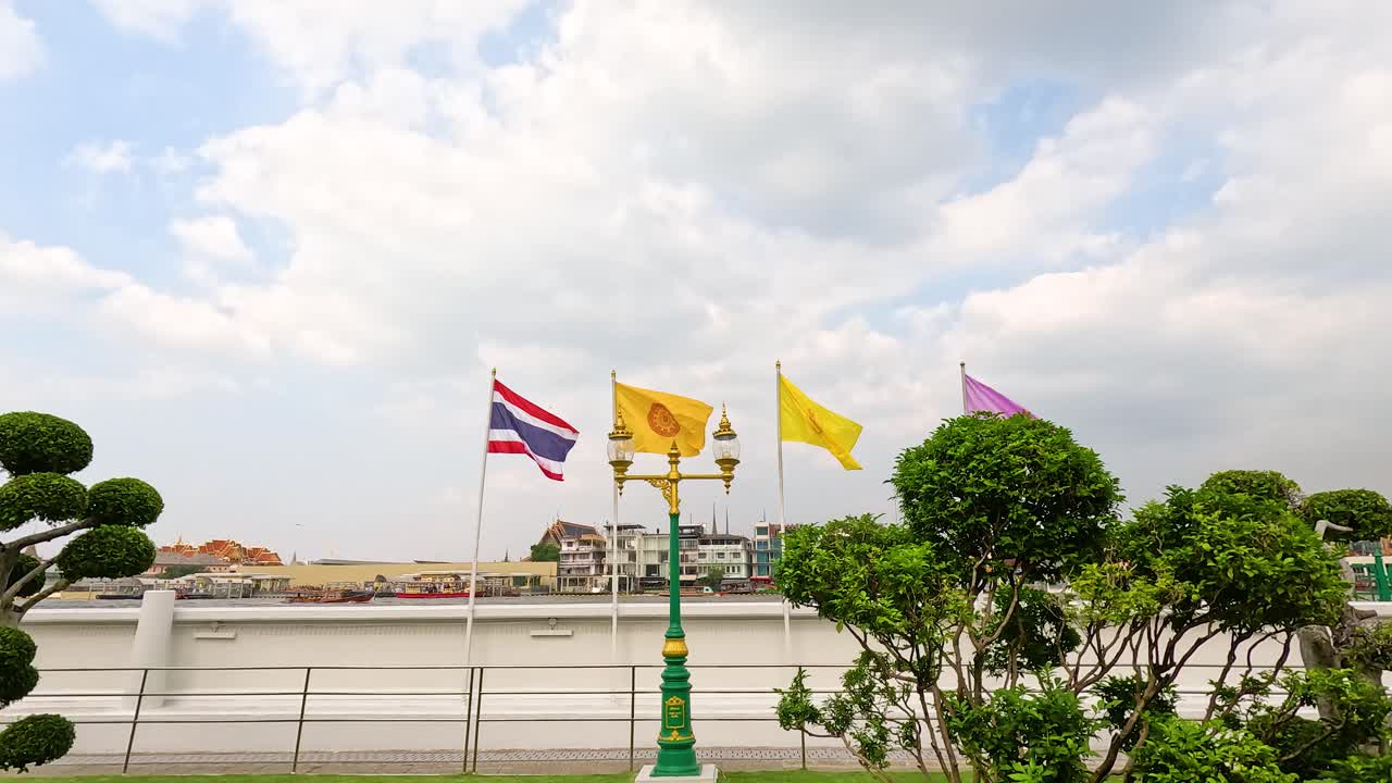 Flags flutter by the riverside near Wat Arun in Bangkok. Bright daylight illuminates the lush greenery and distant cityscape