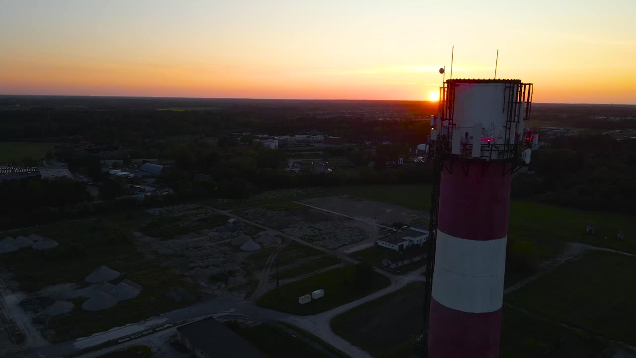 Gorgeous aerial drone footage orbiting and flying around a tall red and white tall smokestack chimney during sunset or sunrise in an urban area with orange sky and sun in the background. High altitude