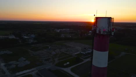 gorgeous aerial drone footage orbiting and flying around a tall red and white tall smokestack chimney during sunset or sunrise in an urban area with orange sky and sun in the background. high altitude