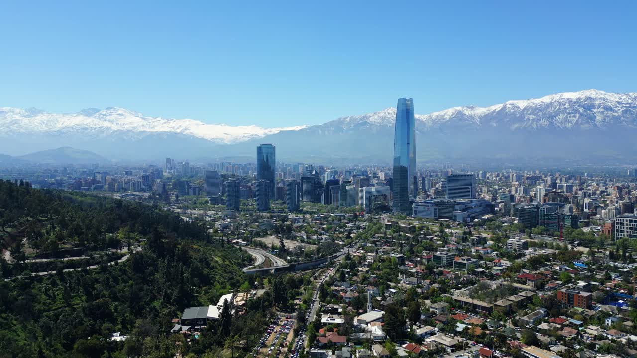 Drone aerial descending past the glass Costanera tower, revealing downtown skyline and snowcapped Andes under clear daytime light. Santiago, Chile