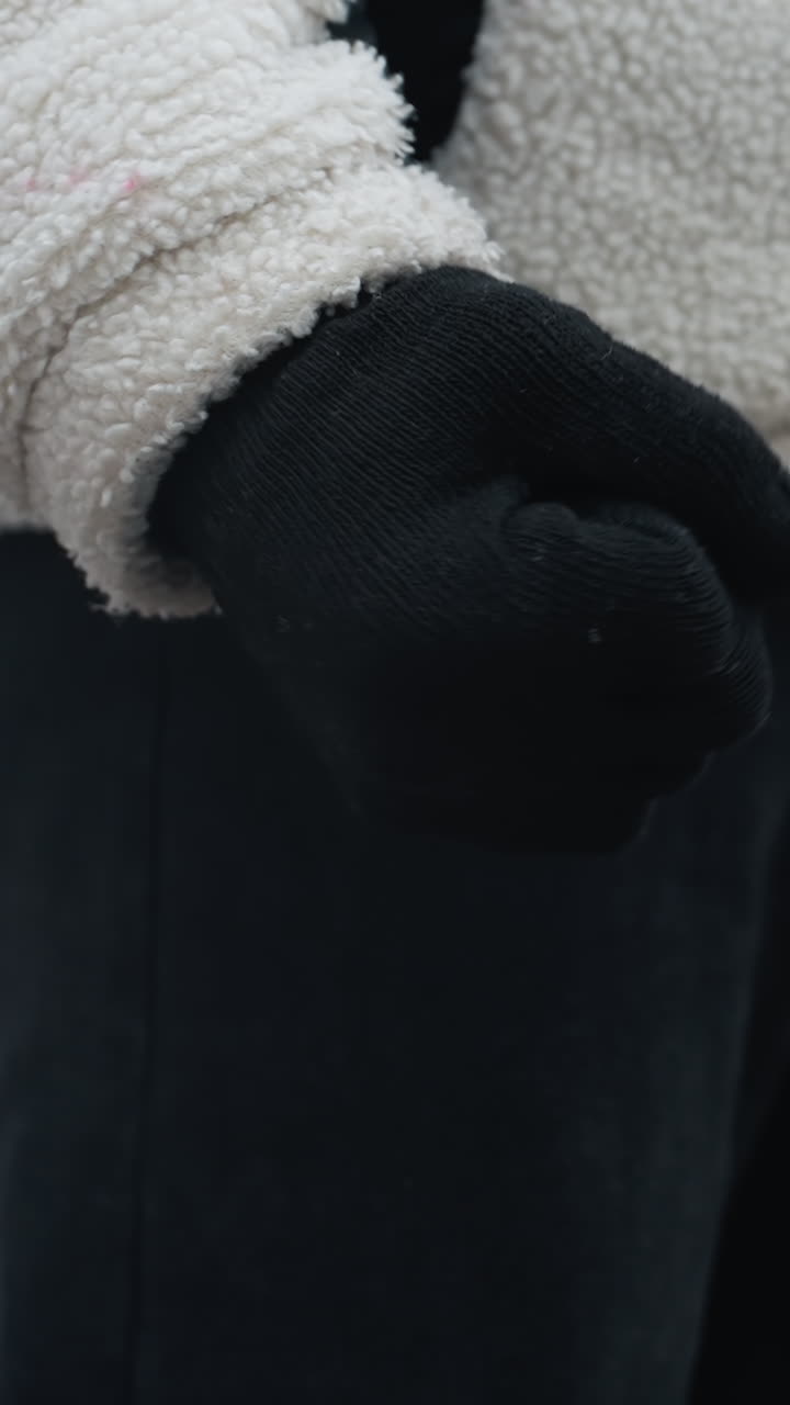 검은 장갑, 겨울 재, 검은 바지를 입은 사람이 손목을 회전시키고, 흐릿한 눈 바탕에 멀리 있는 인물과 지나가는 차를 보여주는 근접적인 측면 시각