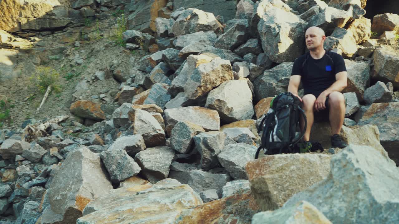 adult tourist with a backpack next to him sitting on a stone and looking at the wonderful nature with rocks around himself