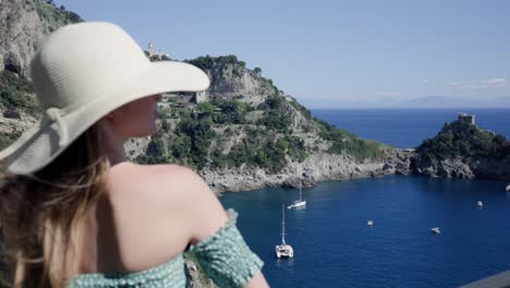 una encantadora mujer caucásica con sombrero y vestido de playa de verano fuera del hombro, de pie en el balcón mirando la vista panorámica del océano con yates amarrando en alta mar en un resort isleño en italia