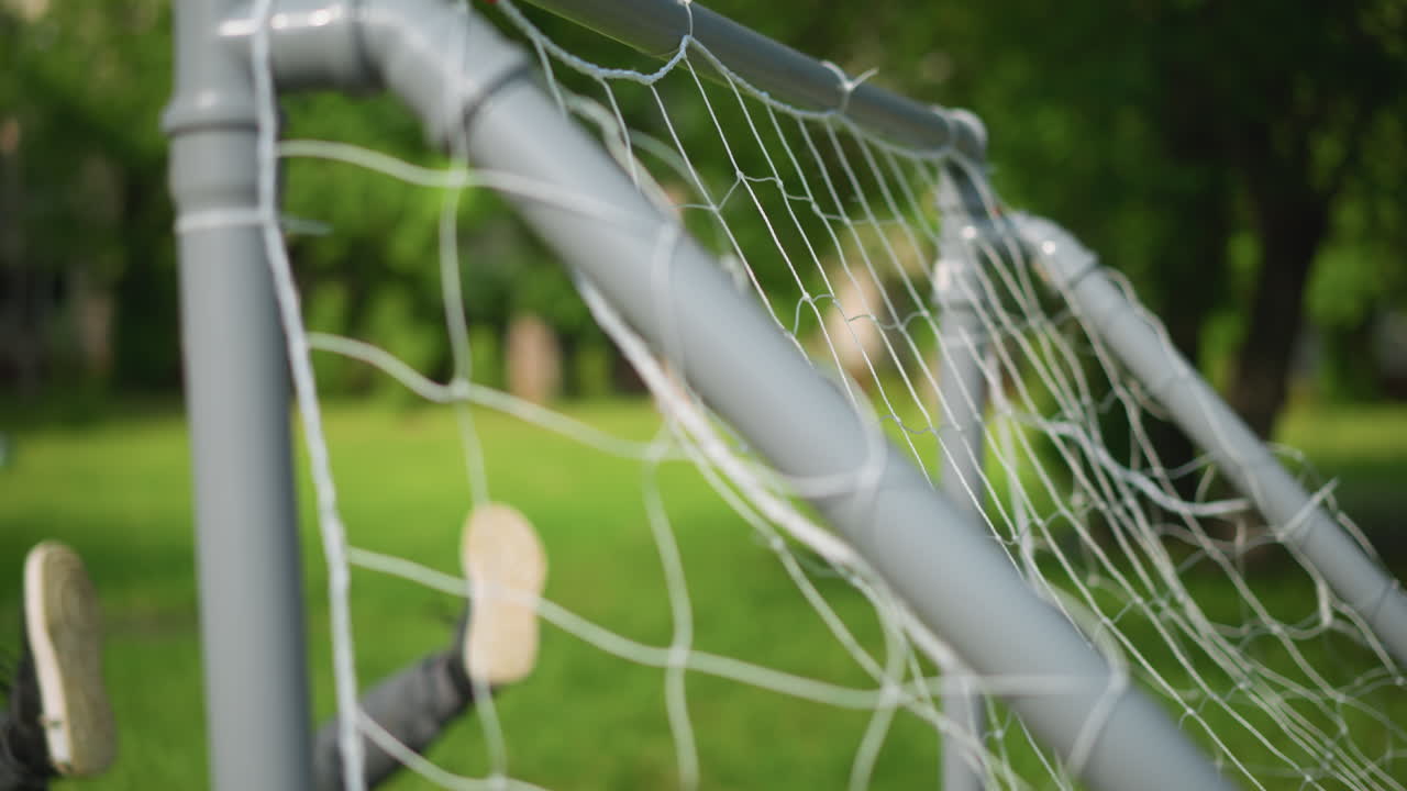 A close-up of a goal post with a ball been shot into the net as the goalkeeper misses and falls, legs up, against a blurred green background