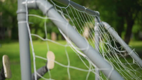 a close-up of a goal post with a ball been shot into the net as the goalkeeper misses and falls, legs up, against a blurred green background