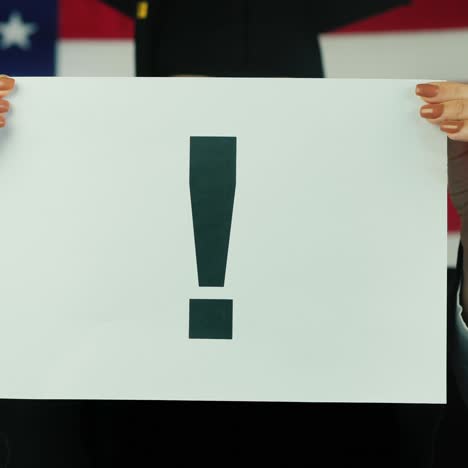 graduado sostiene un cartel con un signo de exclamación contra el fondo de la bandera americana