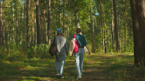 two siblings walking through lush green forest, one wearing blue hair tie and carrying backpack, other with cloth draped over her bag, lifting leg to dodge object on ground