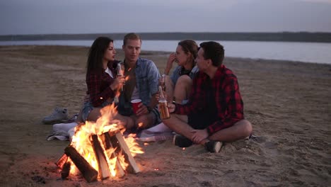 carefree young friends spending time together and drinking beer by bonefire on the beach as the sun begins to set. talking, discussing something