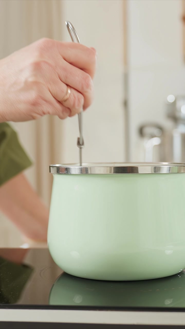 Close up of hand with two rings stirring and adjusting lid. Person wearing olive green top repeatedly lifts slightly. Metallic rimmed open lid cooking vessel sits atop induction