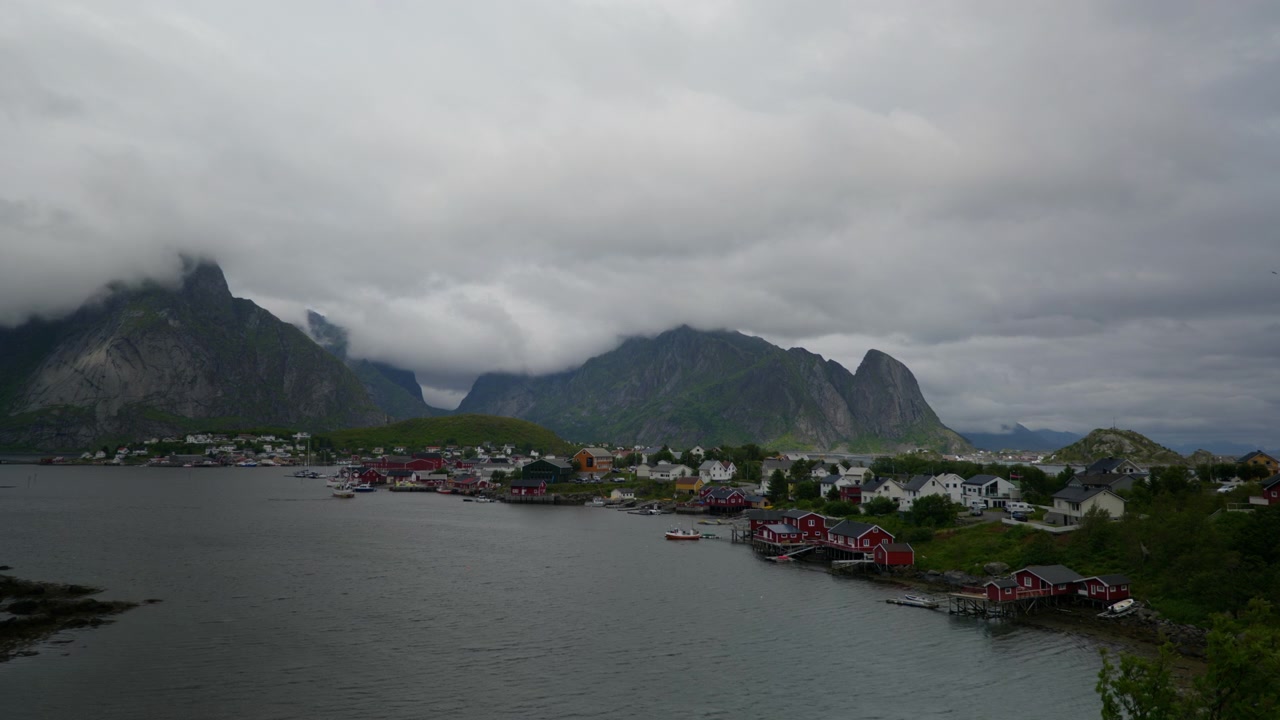 Overskyet himmel hænger over en farverig fiskerby langs en beskyttet bugt, med stejle klippetoppe i baggrunden i Lofoten, Norge