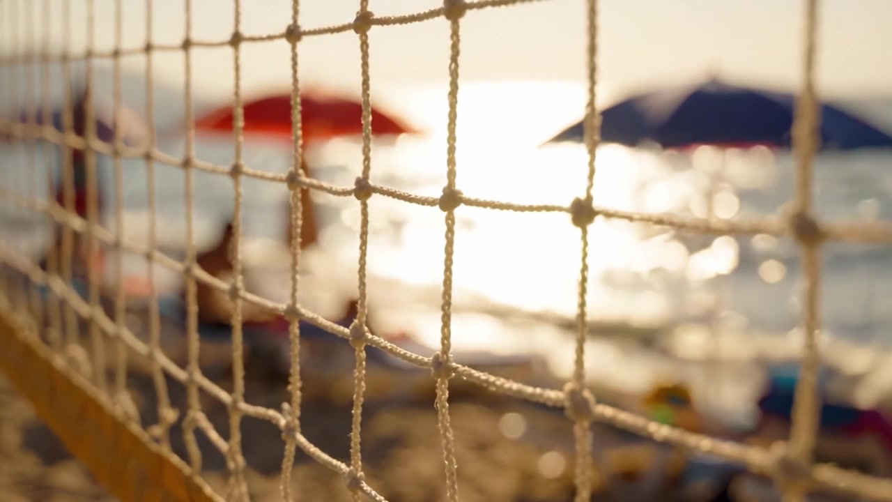 Una tranquilla scena da spiaggia incorniciata da una rete da pallavolo, che mostra ombrelloni vivaci e onde delicate sotto una luce solare dorata.