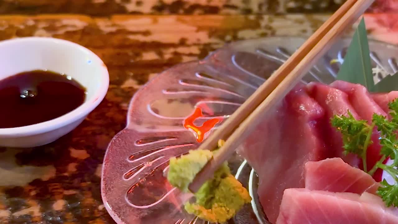 Close-up of chopsticks picking wasabi with sashimi on a decorative plate beside soy sauce.