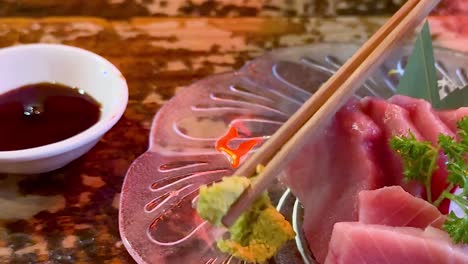 close-up of chopsticks picking wasabi with sashimi on a decorative plate beside soy sauce.