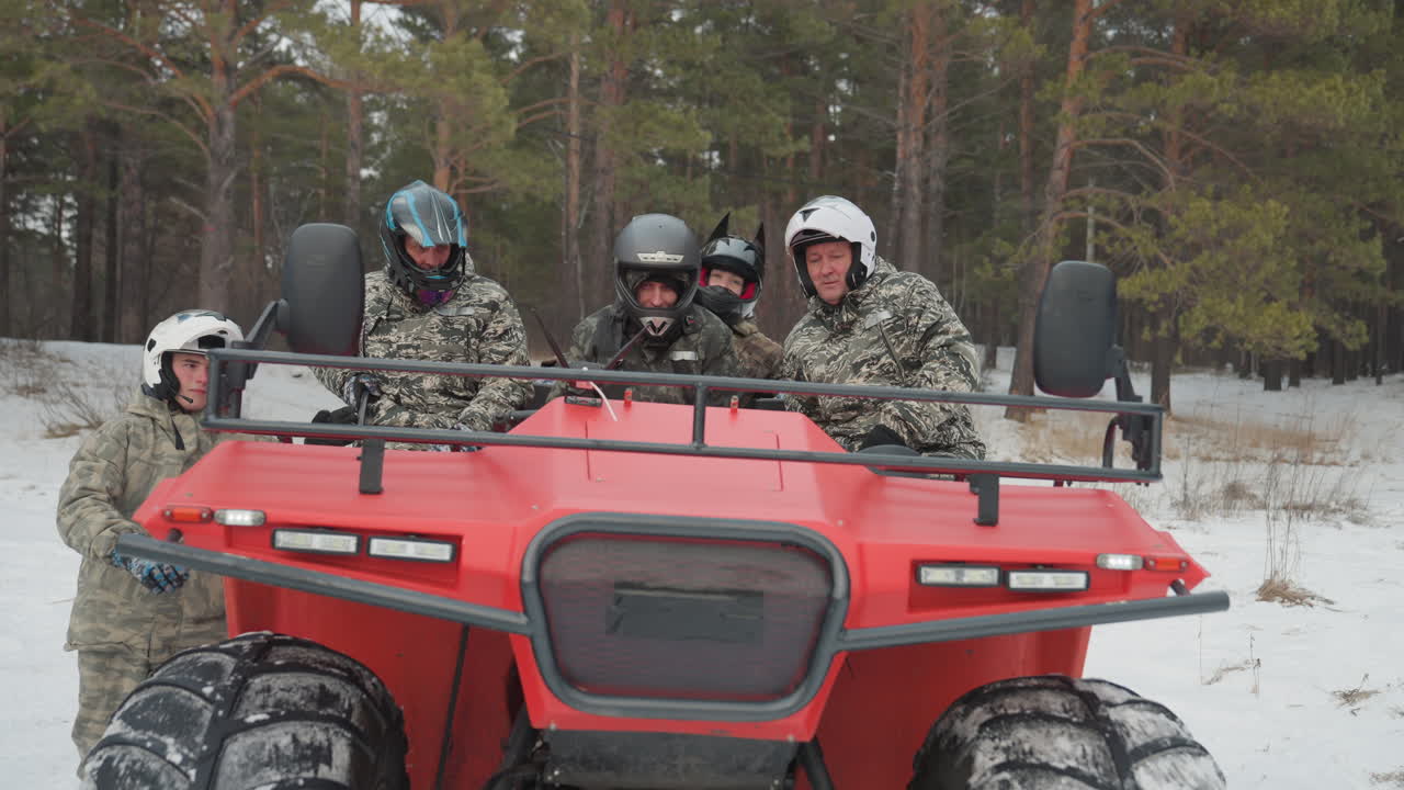 Piloti caucasici che preparano un ATV rosso in una foresta di pini innevata, tre amici con casco e giacche mimetiche che controllano pneumatici e comandi, il conducente che dà brevi istruzioni, passeggeri che regolano l'equipaggiamento, visibile.