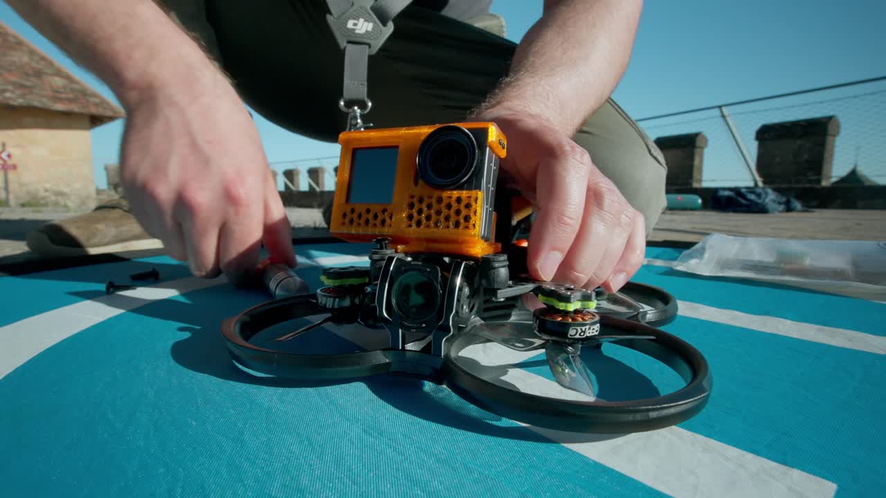 Drone-pilotti kiinnittää oranssin action-kameran elokuvalaatuisen FPV-nelikopterin yläkiinnitykseen asennuksen aikana.