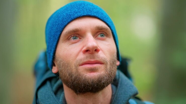 Skjeggete mann med blå lue ser opp i skogen
