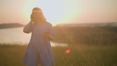 woman in a blue gown enjoys the sunset, holding a black headset and smartphone, gently swaying to the music, highlighted by the sun's warm glow