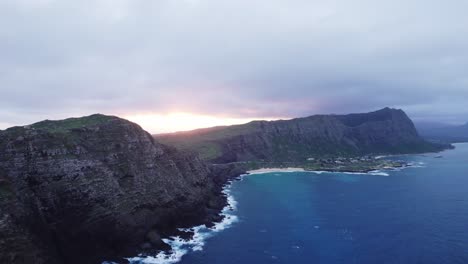 An-aerial-shot-captures-the-breathtaking-view-of-the-sun-setting-behind-the-rugged-cliffs-of-Oahu's-North-Shore,-highlighting-the-contrast-between-the-lush-green-landscape-and-the-vibrant-blue-ocean