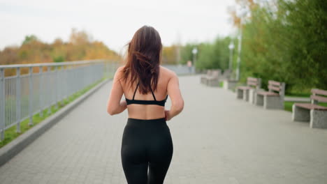 young woman in black singlet and leggings jogging in bright park, background features iron fence, trees, and benches, showcasing an active outdoor lifestyle in sunny, vibrant atmosphere
