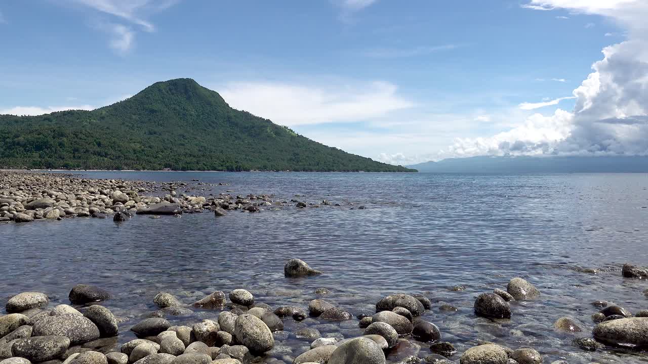 costa de la isla tropical cubierta de rocas con olas que pasan suavemente y una alta montaña cubierta de árboles en el fondo bajo un cielo azul brillante y nublado