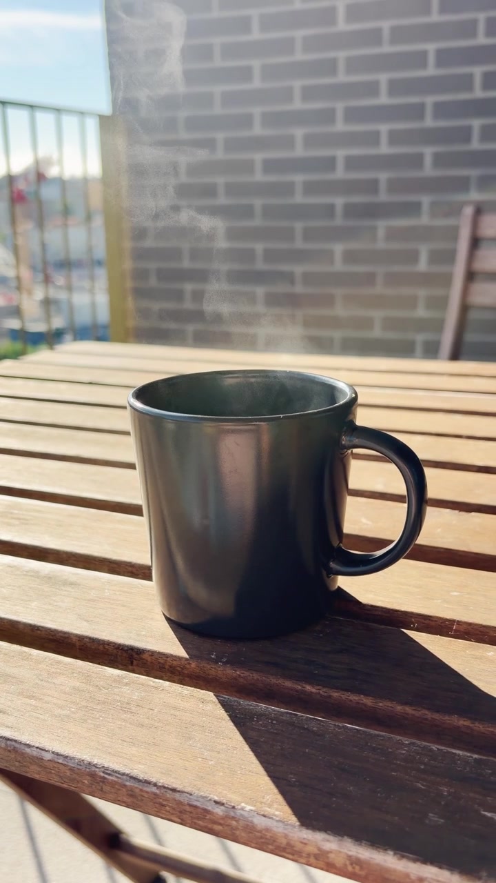 Person's hand placing a black mug of hot, steaming coffee on a sunlit wooden table on a balcony, creating a tranquil and cozy moment for a perfect start to the day or a relaxing break