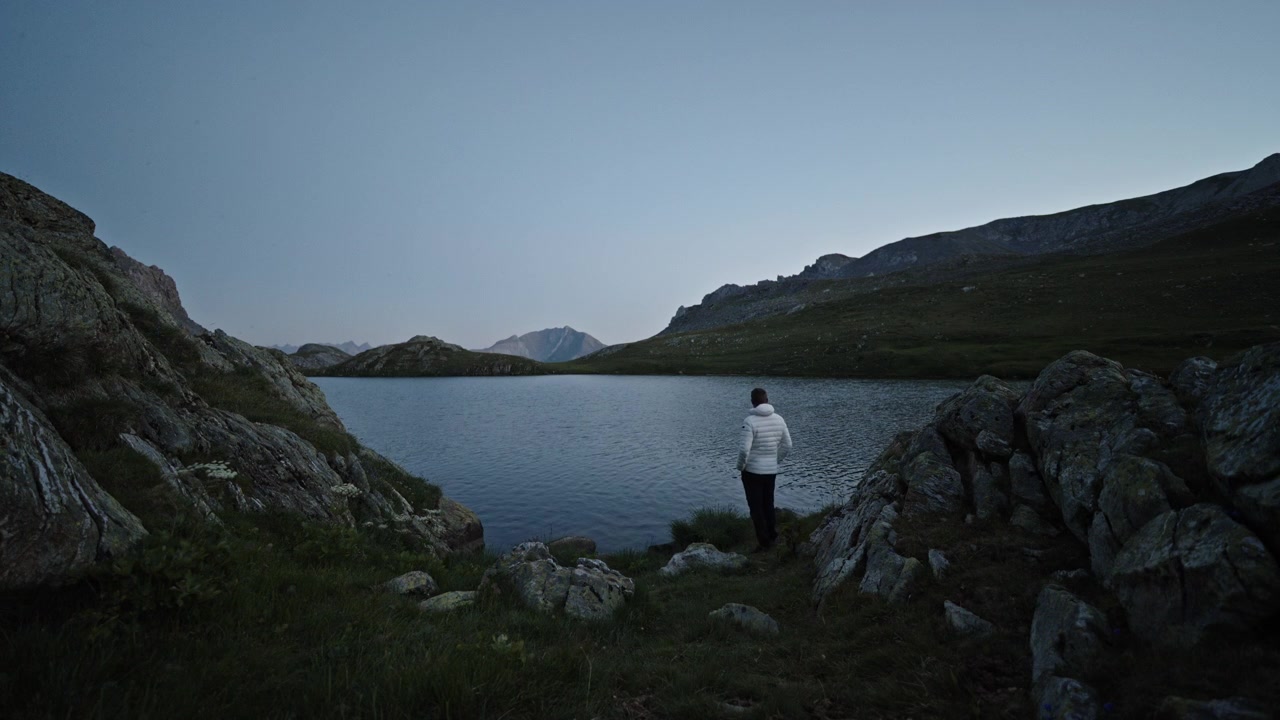 Un escursionista solitario con una giacca bianca sta in silenzio vicino a un lago alpino italiano al tramonto, incorniciato da massi e creste distanti