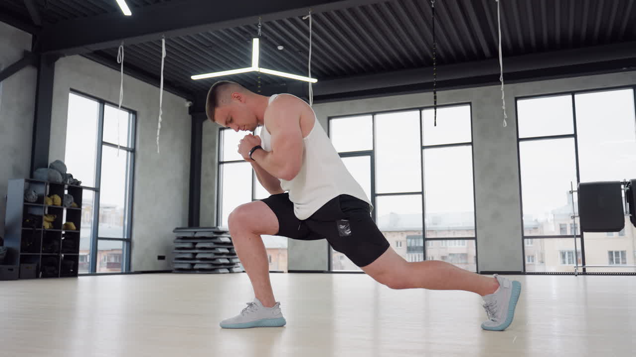 Gym rat performing side lunge stretch in bright studio, clasping hands and extending leg behind on wooden floor, wearing white tank top and black shorts, large windows and hanging straps overhead