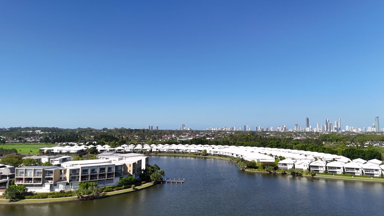 Drone pans from riverside trees to modern lakeside residences with distant city skyline under sunlight