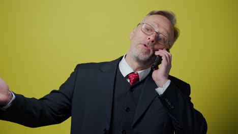 nervous middle aged businessman with a cellphone. mature man in dark suit talking on the phone and giving instructions to somebody on yellow background.