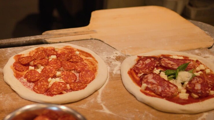 Lo chef aggiunge pepperoni e olio d'oliva a pizze crude su un bancone di legno infarinato
