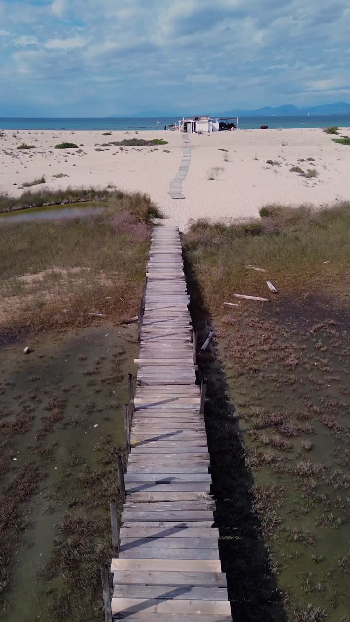 Ripresa verticale con drone 9:16 in volo sopra un lungo ponte rustico in legno verso una baracca da spiaggia remota a Lefkada, Grecia. Scenario cinematografico di zone umide costiere, ideale per reel di social media e TikTok.