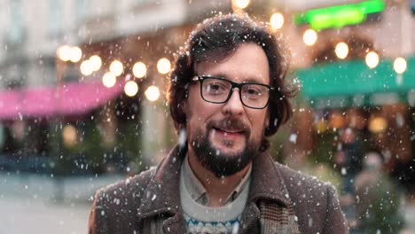 close-up view of caucasian man wearing eyeglasses looking at camera on the street while it‚äôs snowing in christmas