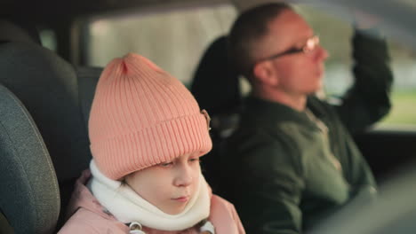 a close-up shot of a young girl wearing a pink beanie and warm jacket, sitting quietly in a car, looking pensive, with a man, possibly her father, blurred in the background