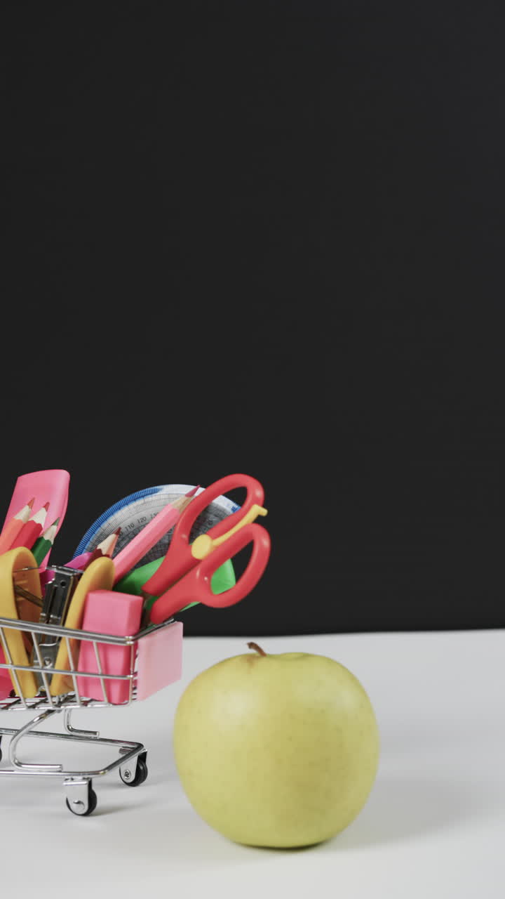 Vertical video of shopping trolley with school materials, apple and copy space on grey background