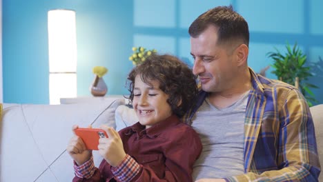 Portrait-of-happy-father-and-son-having-fun-using-smartphone-on-sofa-in-living-room.