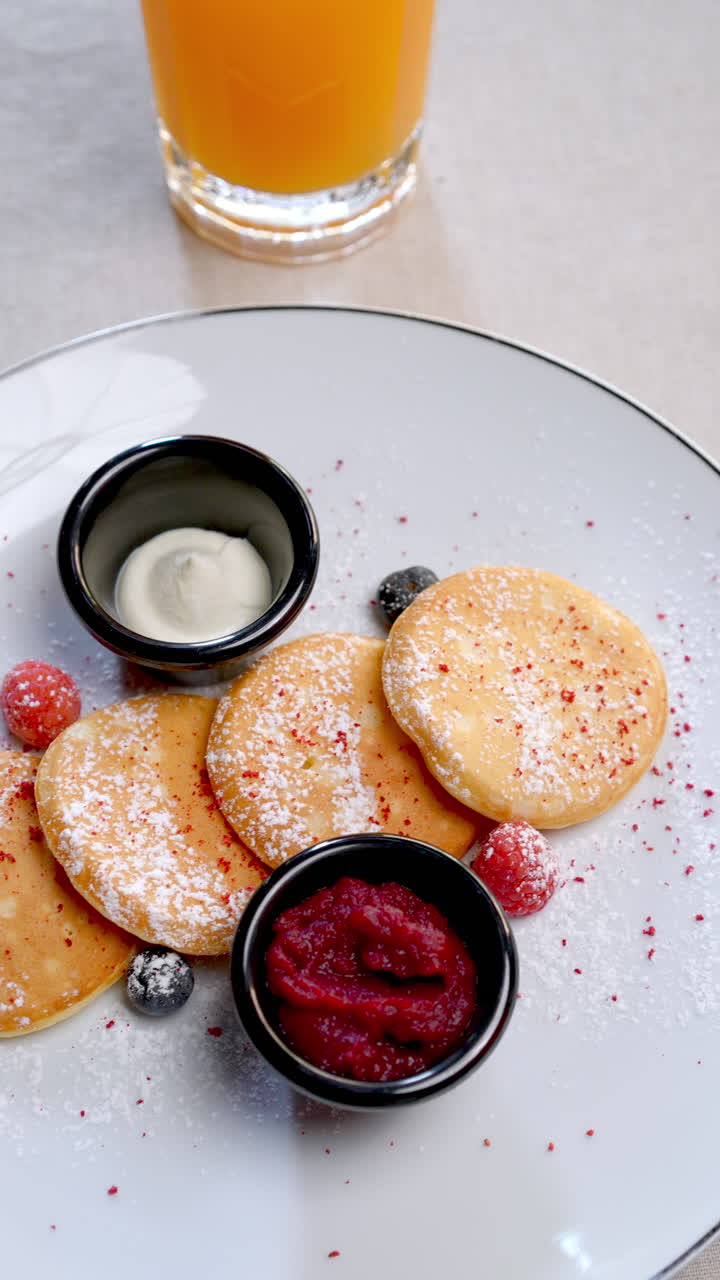 Small pancakes are served on a white plate with fresh berries, cream and jam, accompanied by a glass of orange juice, composing a healthy and delicious breakfast
