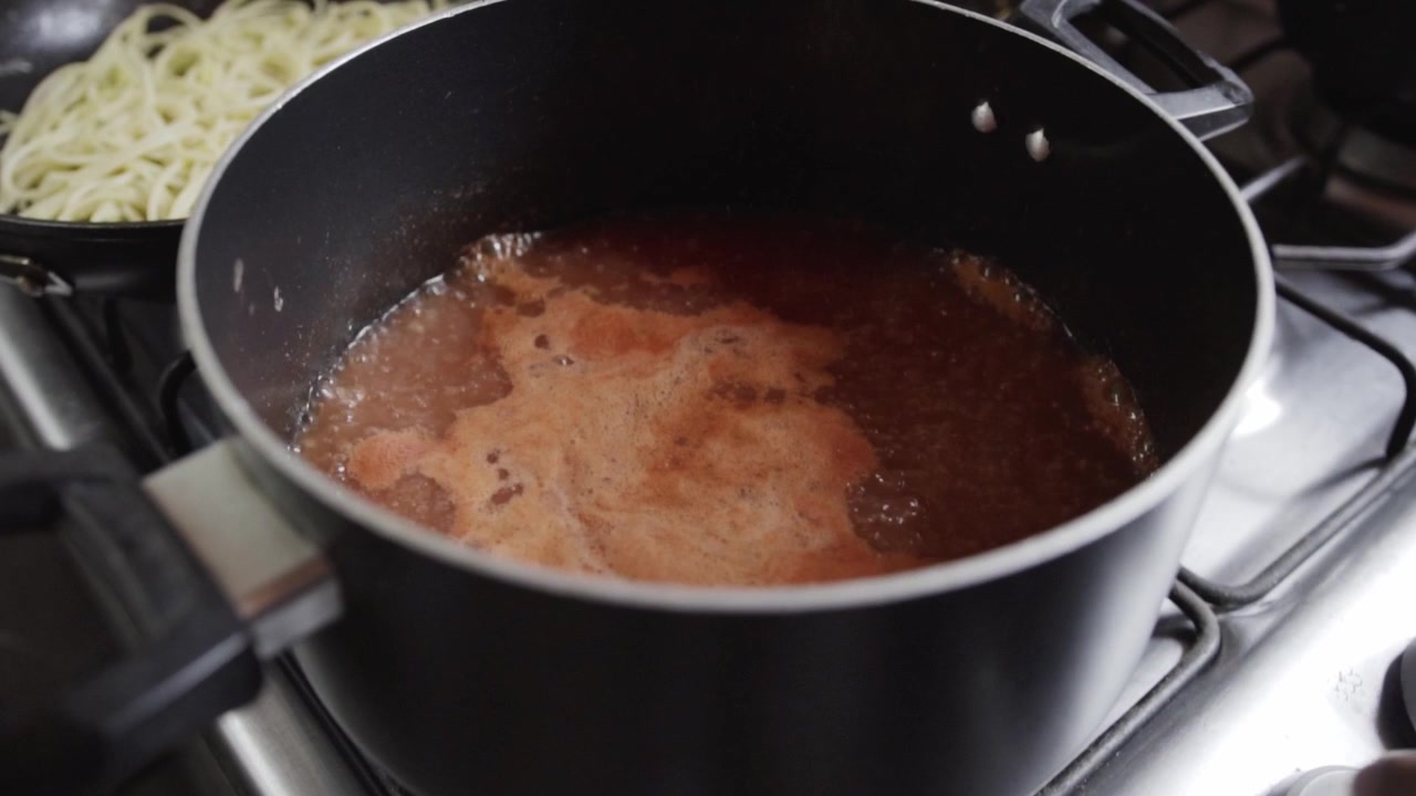 Closeup of tomato sauce simmering in a cooking pot on a gas stove with bubbling liquid and pasta ready for serving during home meal preparation in a kitchen