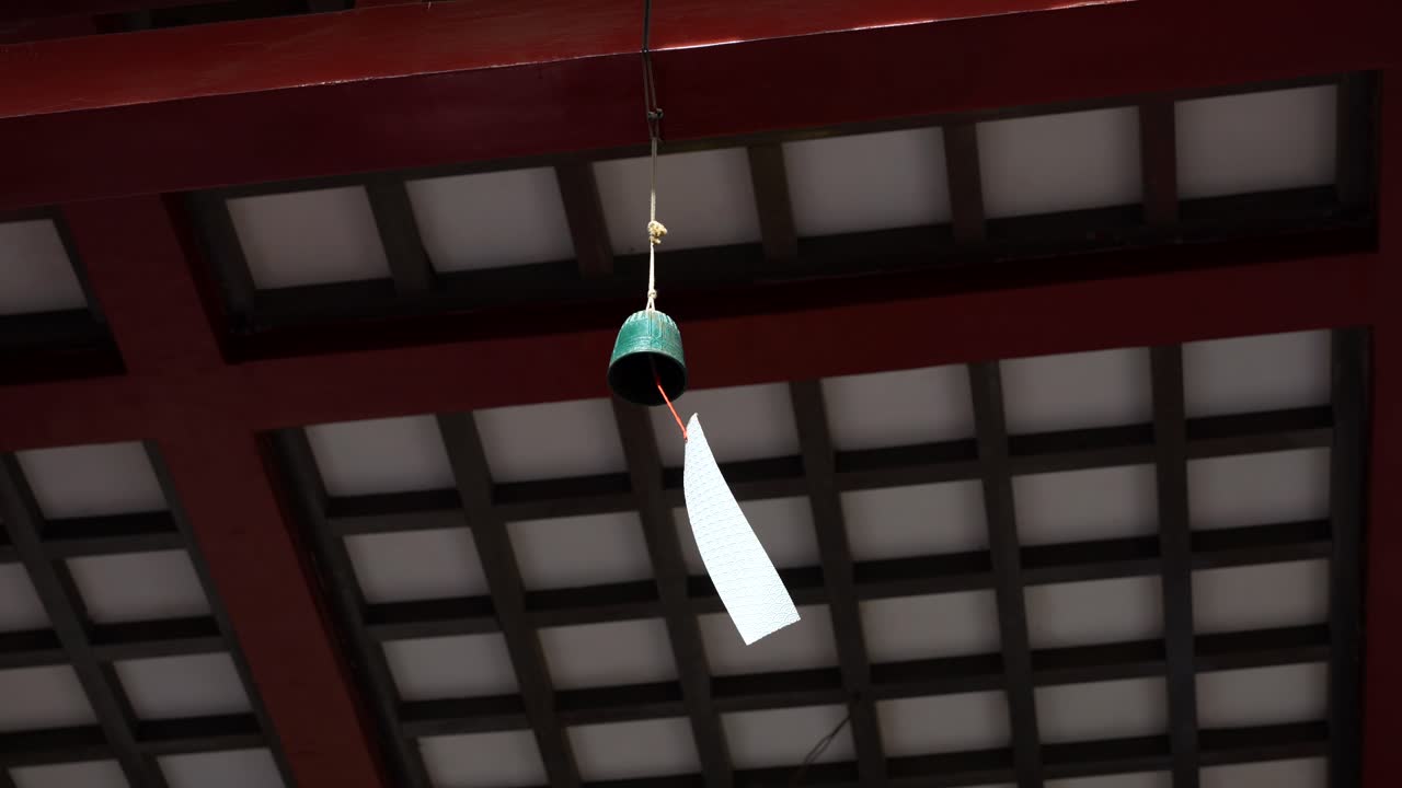 Small green bell wind chime with white paper strip hangs from red wooden beam ceiling. Low angle