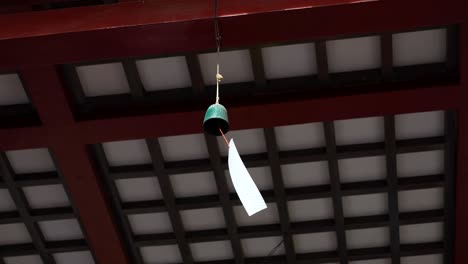small green bell wind chime with white paper strip hangs from red wooden beam ceiling. low angle