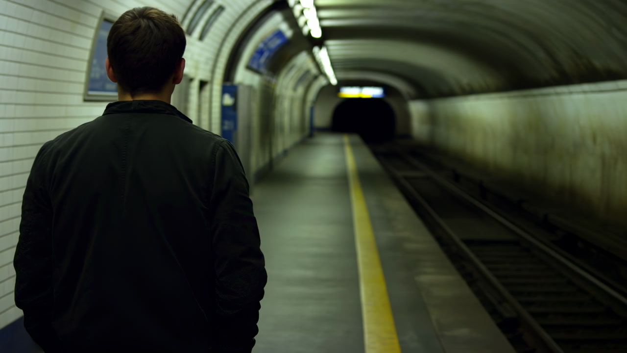 Man Standing On Empty Subway Platform, Back To Camera, Looking Into Dark Tunnel With Moody Fluorescent Lights,