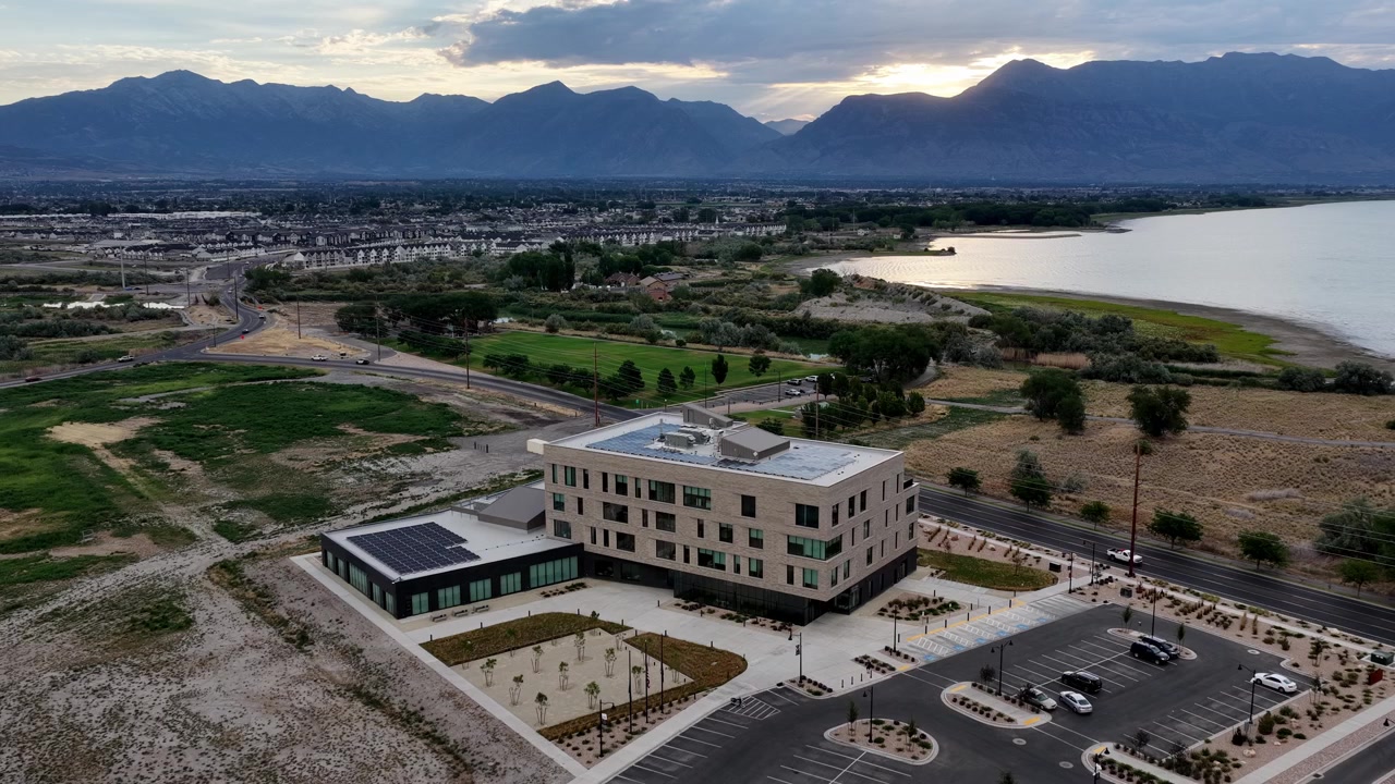 Letecké záběry dronu se stahují zpět nad jezerem Utah a Saratoga Springs, odhalující moderní solární kancelářskou budovu, svěží pole, rozlehlé parkoviště a malebné horské pozadí při východu slunce