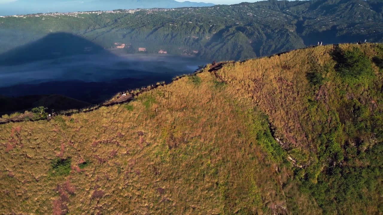 Ampio paesaggio montano con escursionisti che ammirano un'enorme caldera vulcanica e altopiani aspri nella morbida luce del mattino sopra un vasto terreno craterico, riprese aeree con drone.
