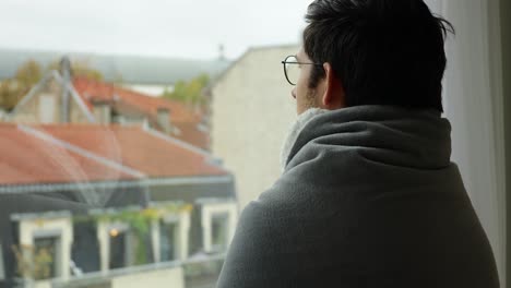 young man wearing hot blanket and looking through a window during overcast day