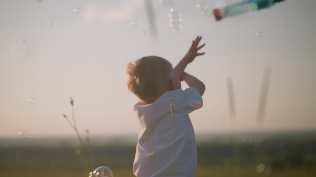 A joyful little boy in a white shirt stands in a grassy field with his hands raised, eagerly touching bubbles floating around him in the warm sunlight, capturing a moment of pure childhood happiness