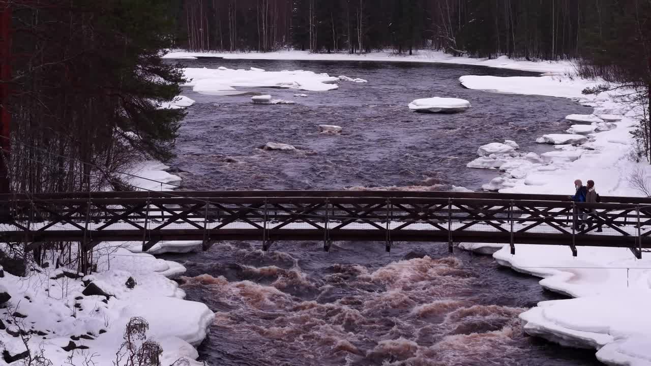 Pari kävelee sillalla joen yläpuolella, jonka punertavanruskea sävy virtaa läpi mäntymetsämaiseman taustalla, Suomi, ilmakuva